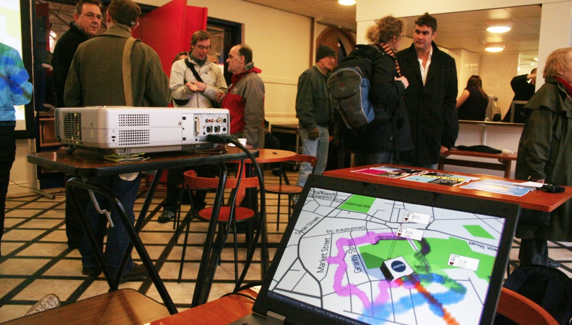 People milling about in a room. There is a laptop open on a table, which shows a map overmaked with pink, blue and orange tracks showing where people have walked.