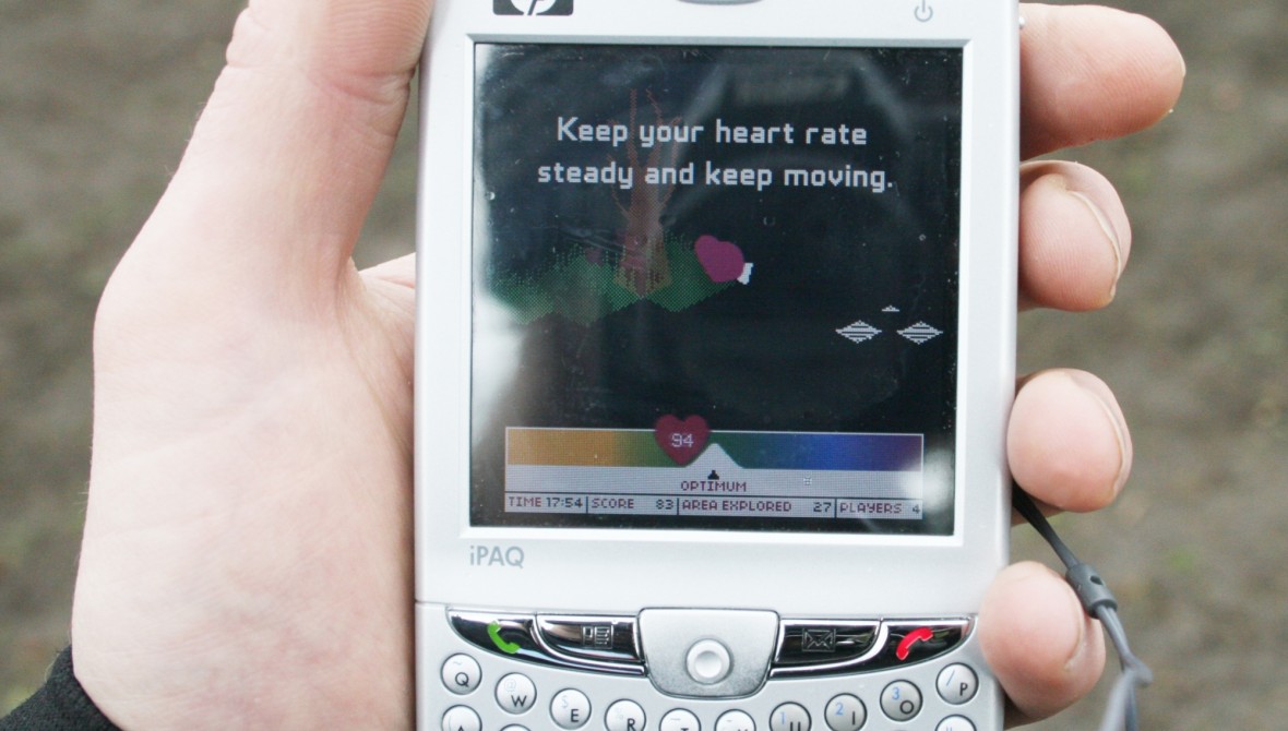 A hand holding a mobile phone. The screen shows a heart icon by a tree and the text 'Keep your heart rate steady and keep moving.' A bar at the bottom displays a current heart rate.