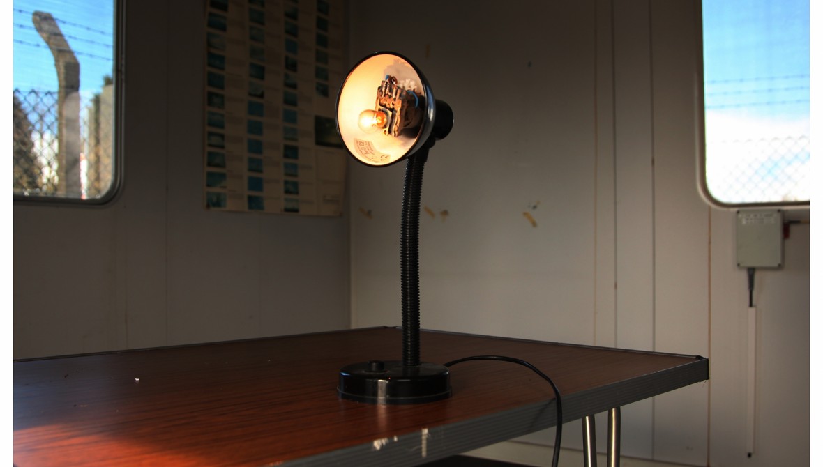A lamp on a table in the corner of a portakabin room. Attached to the bulb is a sensor.