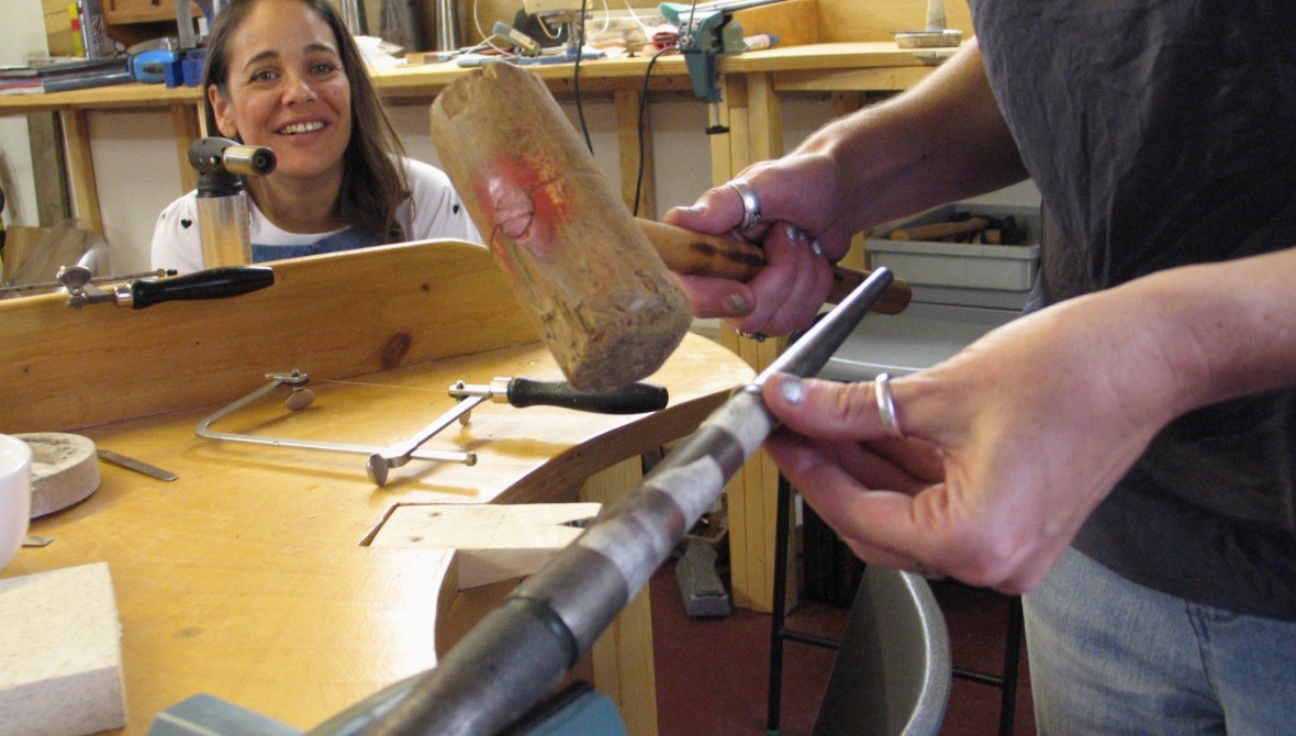 A woman peers at a person (head out of shot) holding a large wooden mallet and fixing a rod in a vice.