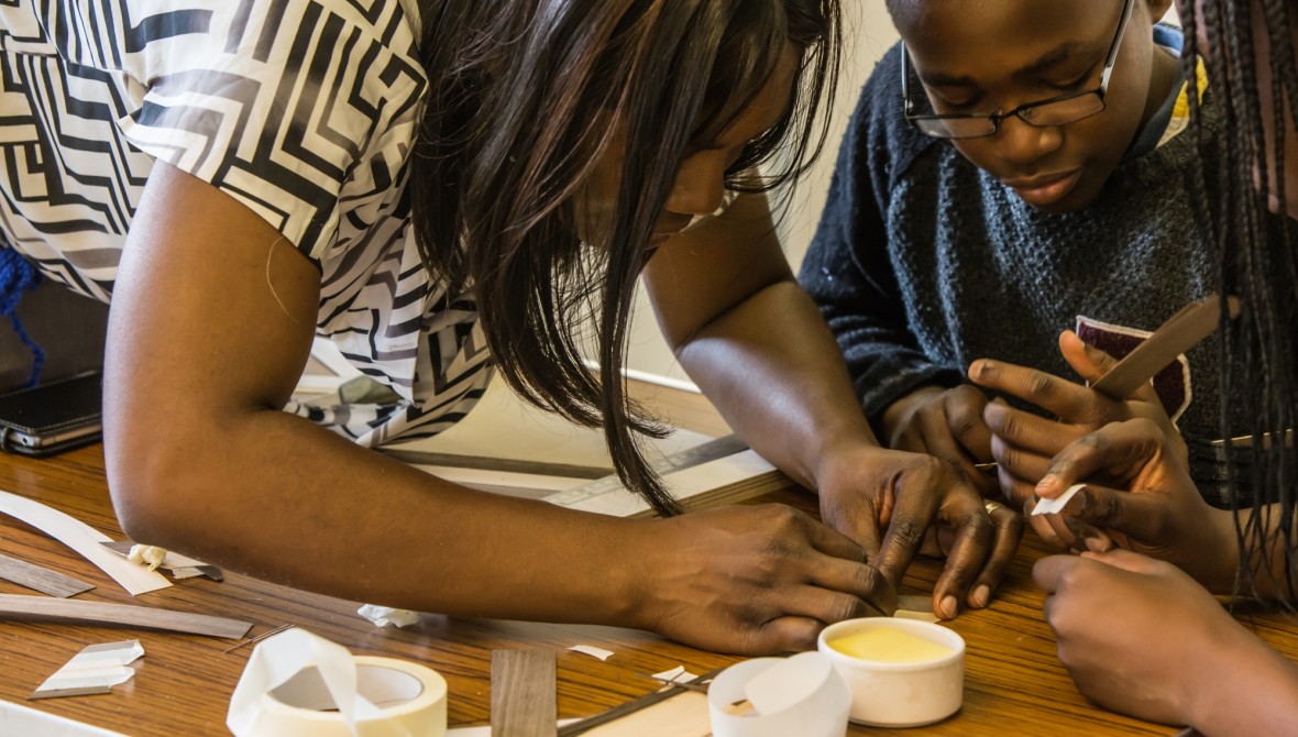 A woman and two children bent over a table covered in craft materials, focussing on something small and fiddly (out of shot).