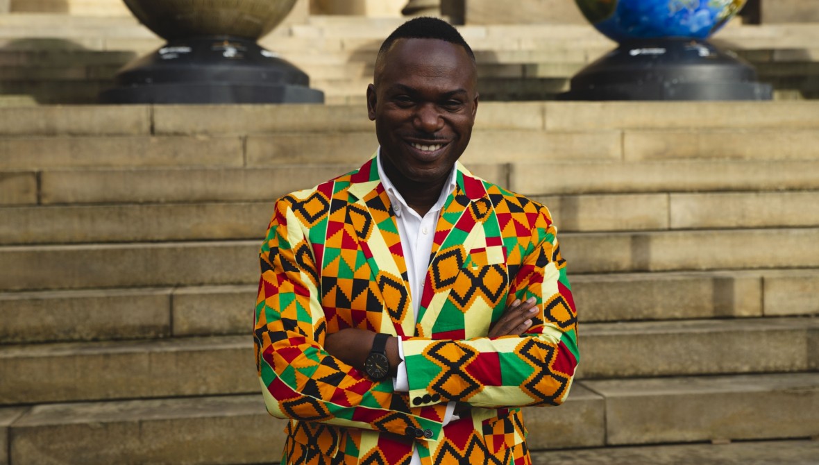 Jason Allen-Paisant, a black man with short black hair in his 30s-40s, stands with his arms crossed, in front of stone steps. He is wearing a bright multi-coloured suit jacket and white buttoned shirt.