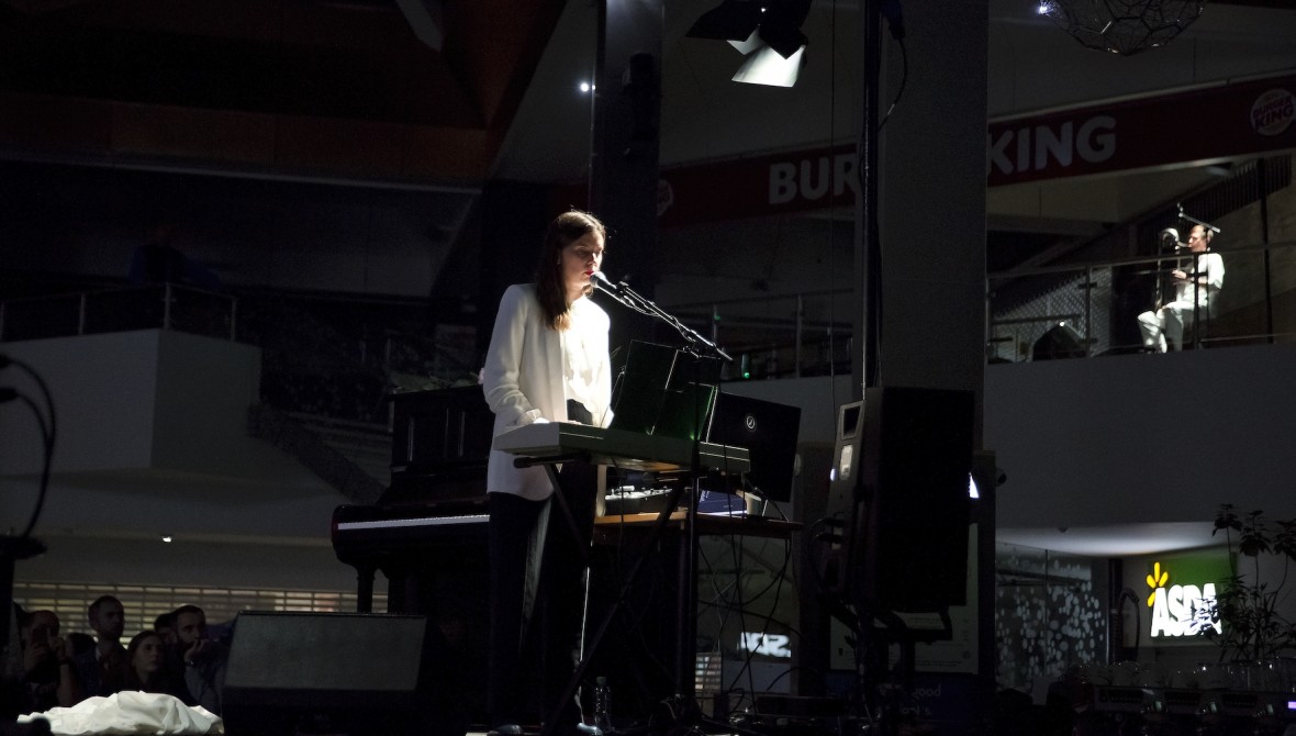 Alice Theobald speaks into a microphone attached to a keyboard on a stage in a dimly lit shopping centre with Burger King and Asda in the background