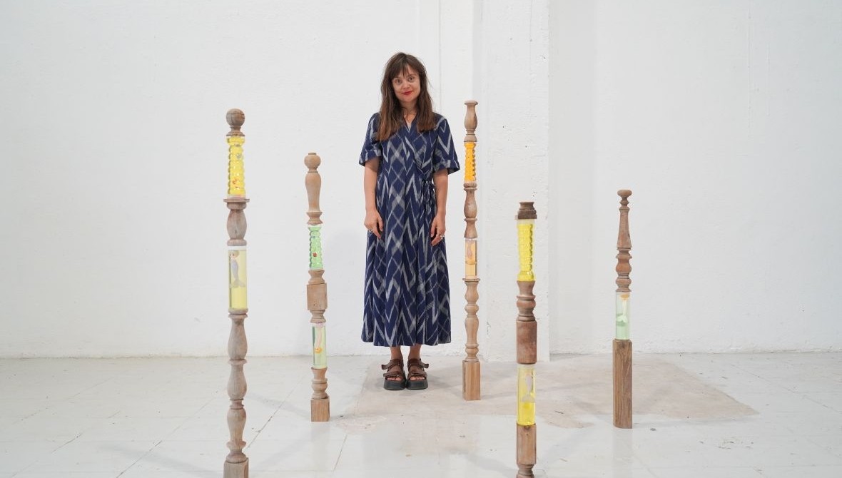 White female artist with long dark brown hair and red lipstick wearing a blue patterned dress, standing in an exhibition space surrounded by 5 structures that look like balusters or spindles made of wood with sections that are clear and appear to be fille