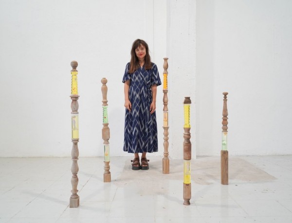 White female artist with long dark brown hair and red lipstick wearing a blue patterned dress, standing in an exhibition space surrounded by 5 structures that look like balusters or spindles made of wood with sections that are clear and appear to be fille